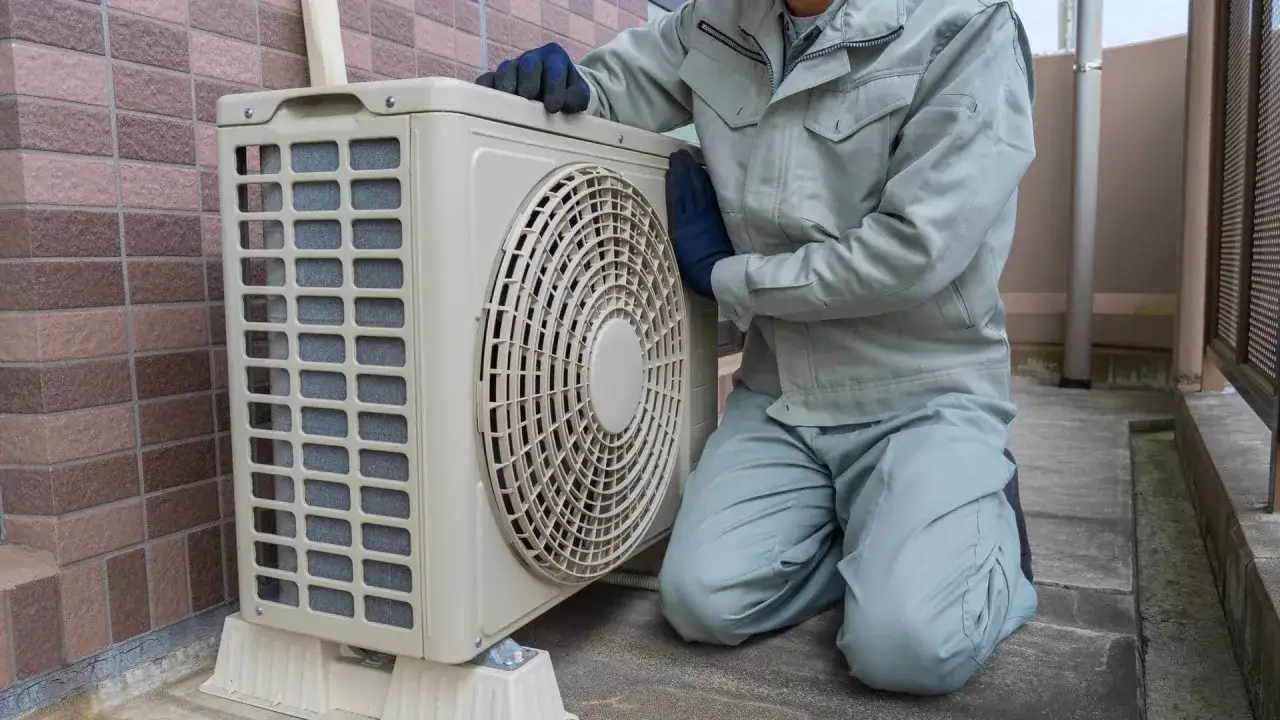 Technik sprawdza klimatyzację w bloku. Urządzenie stoi na zewnątrz, obok ceglanej ściany.