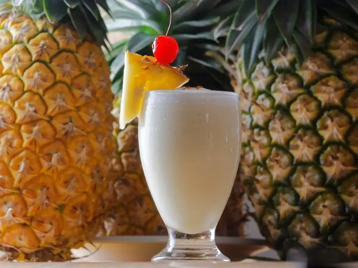 Pina Colada na mleczku kokosowym czy na śmietance? Orzeźwiający drink z ananasem i wisienką, otoczony dojrzałymi owocami.