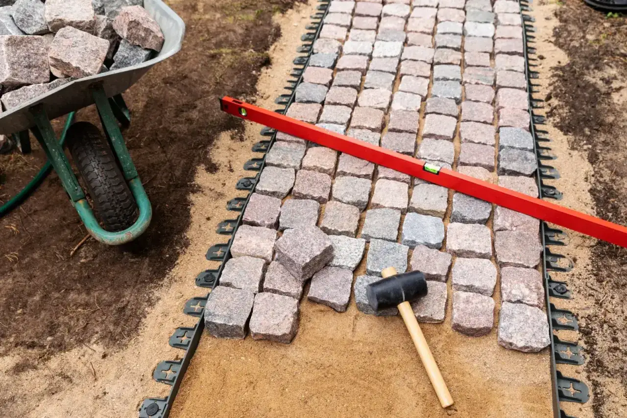 Układanie kostki brukowej na podbudowie z piasku, z użyciem poziomicy i gumowego młotka. W tle taczka z kamieniami.