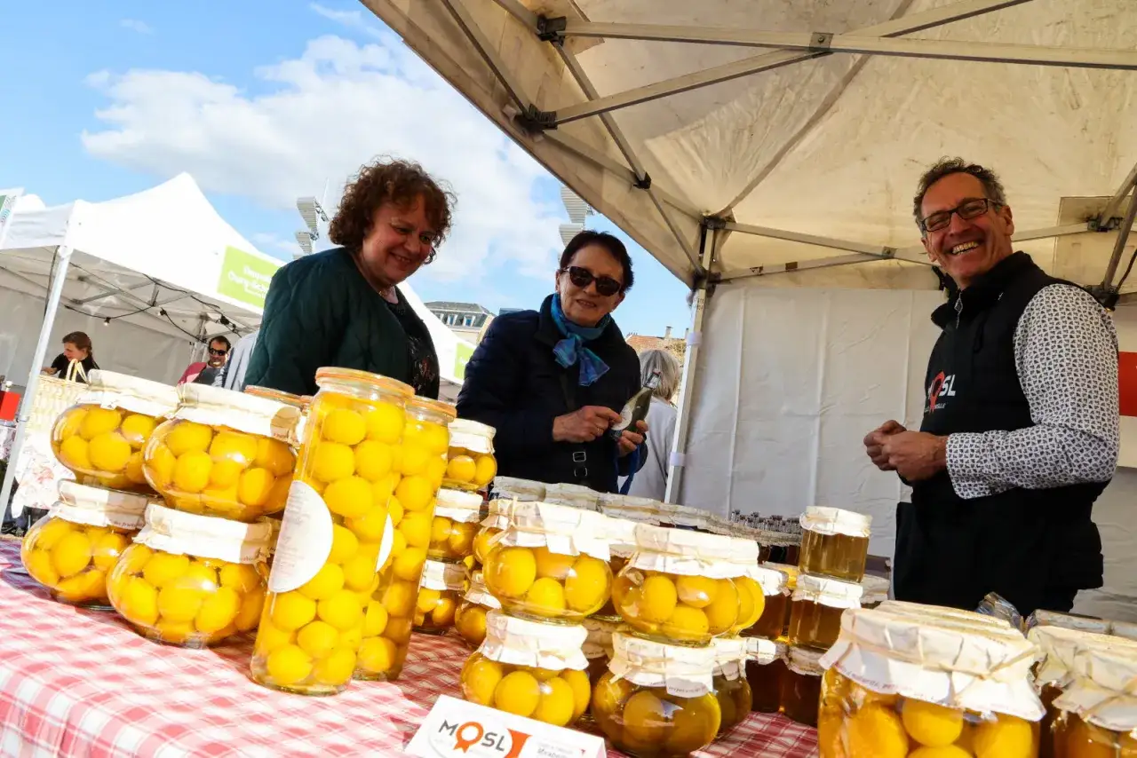 Des mirabelles en bocaux, spécialités de Metz, présentées sur un marché. Trois personnes souriantes discutent devant les étals.