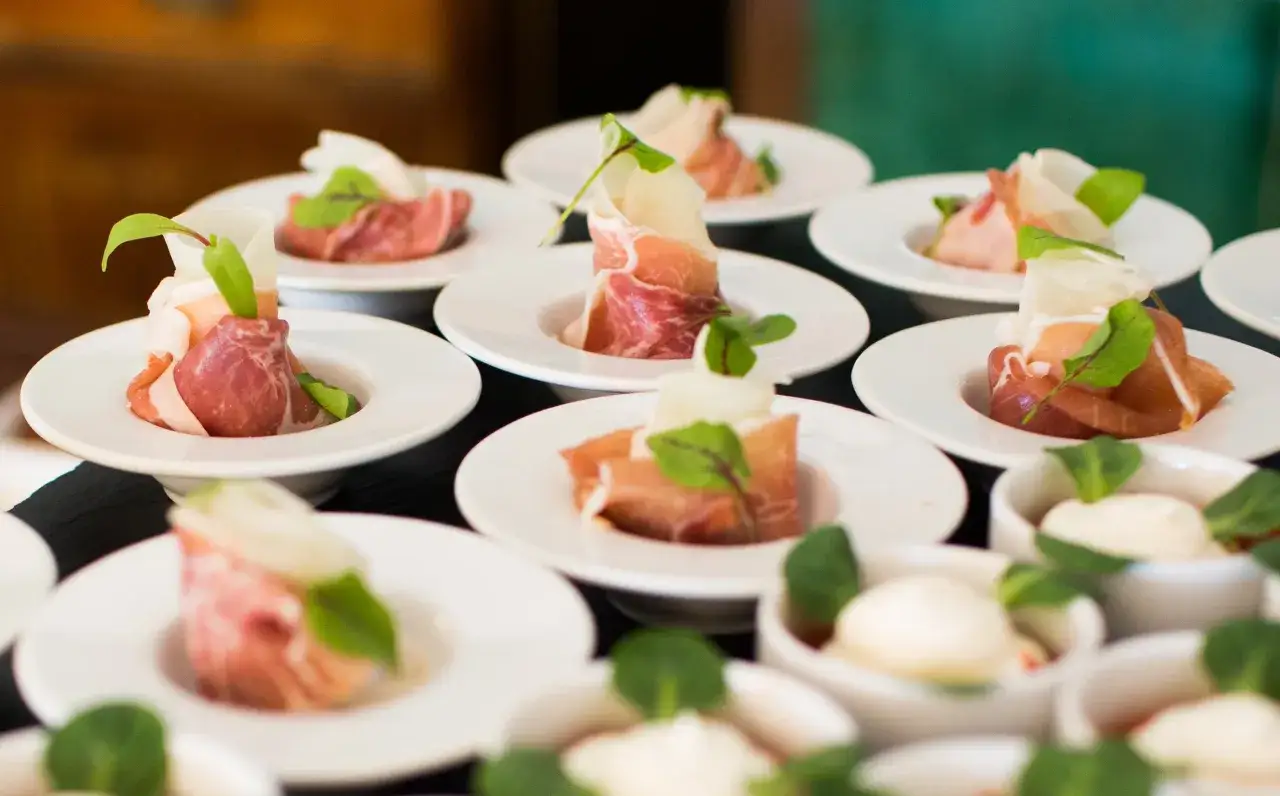 Eleganckie przekąski na białych talerzykach, zawinięte w szynkę parmeńską z listkami ziół. To właśnie catering, który zachwyca smakiem i wyglądem.
