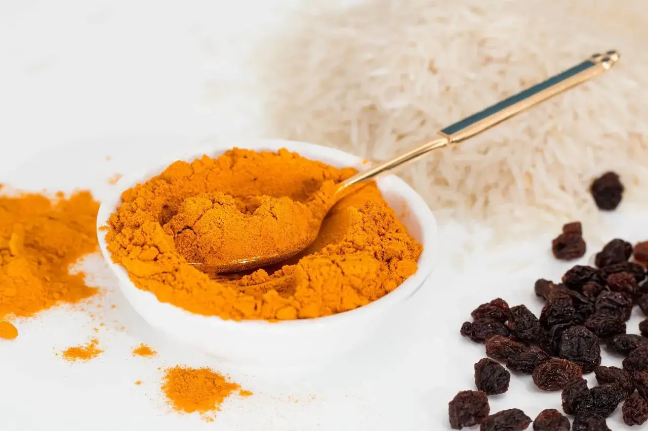 Miseczka pełna intensywnej przyprawy curry, obok ryż i rodzynki. Złota łyżeczka zanurzona w aromatycznym proszku.