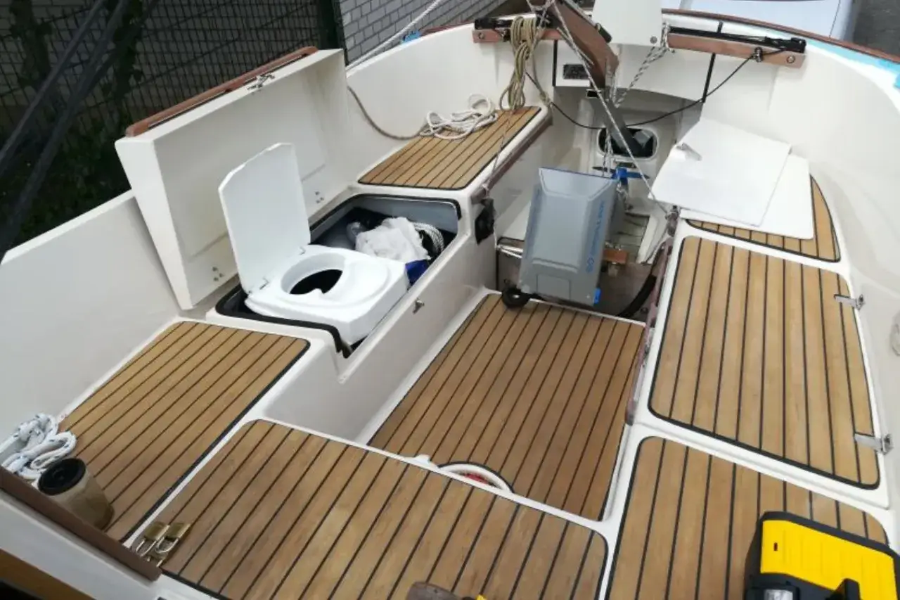 Ein weißes **Segelboot Toilette** mit offenem Deckel, daneben ein grauer Tank. Holzdecks und weiße Wände.