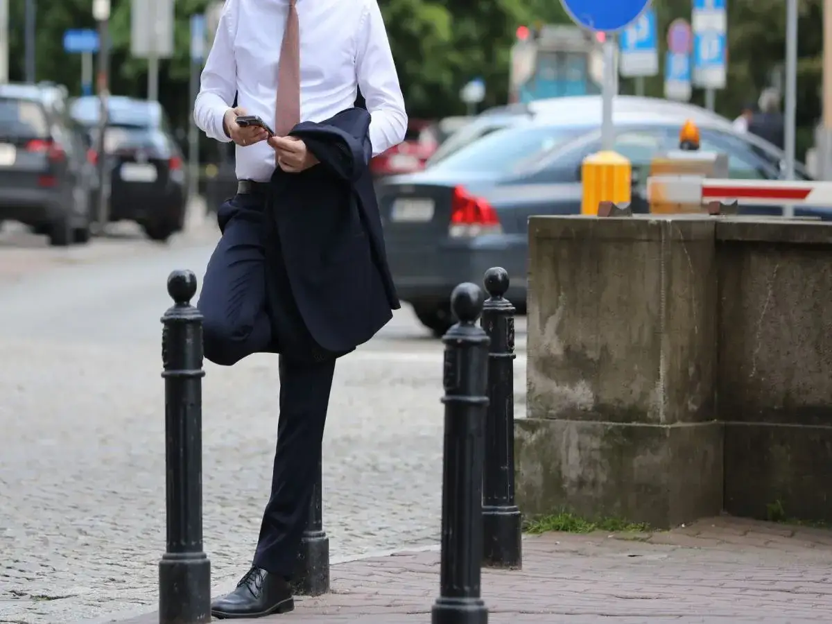 Mężczyzna w garniturze trzyma telefon i marynarkę, opierając się o słupek.