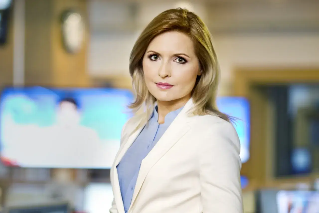 Agnieszka Gozdyra w białej marynarce i niebieskiej koszuli, w tle ekrany telewizyjne. Czy Agnieszka Gozdyra ma dzieci? Tego nie widać na zdjęciu.