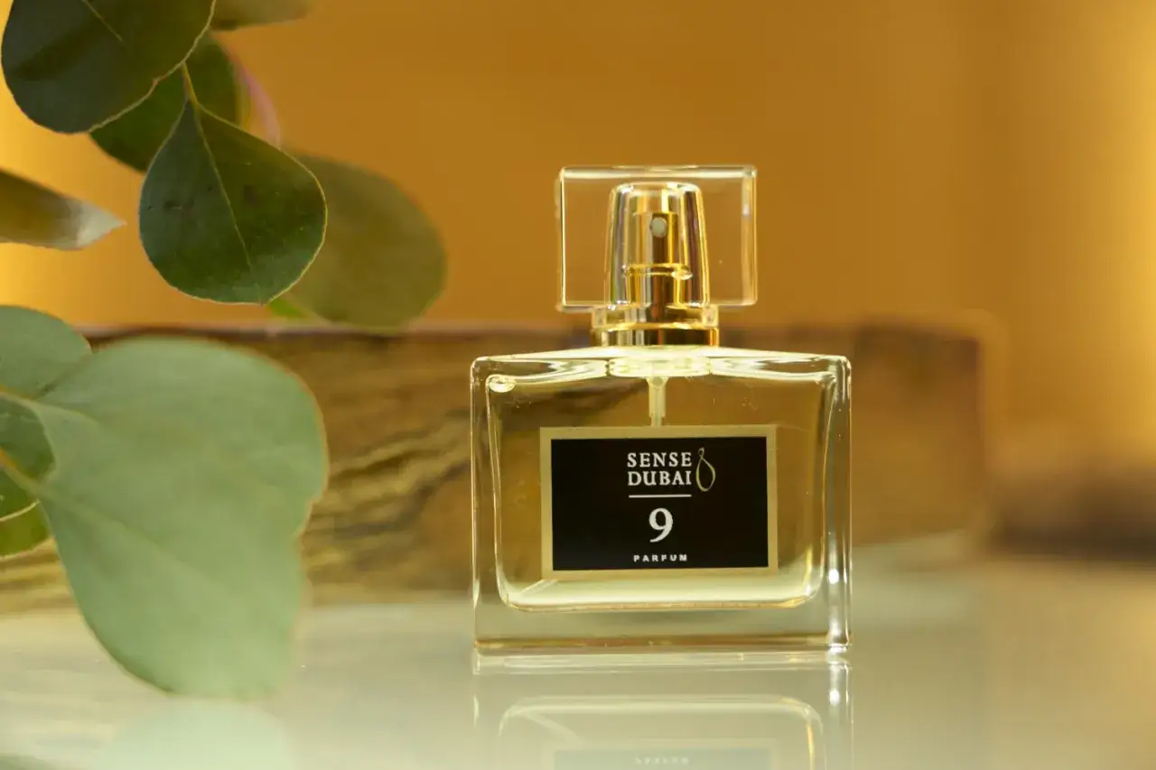 Eleganckie perfumy damskie z Dubaju, Sense Dubai 9 Parfum, w szklanym flakonie z złotymi detalami, otoczone zielonymi liśćmi.