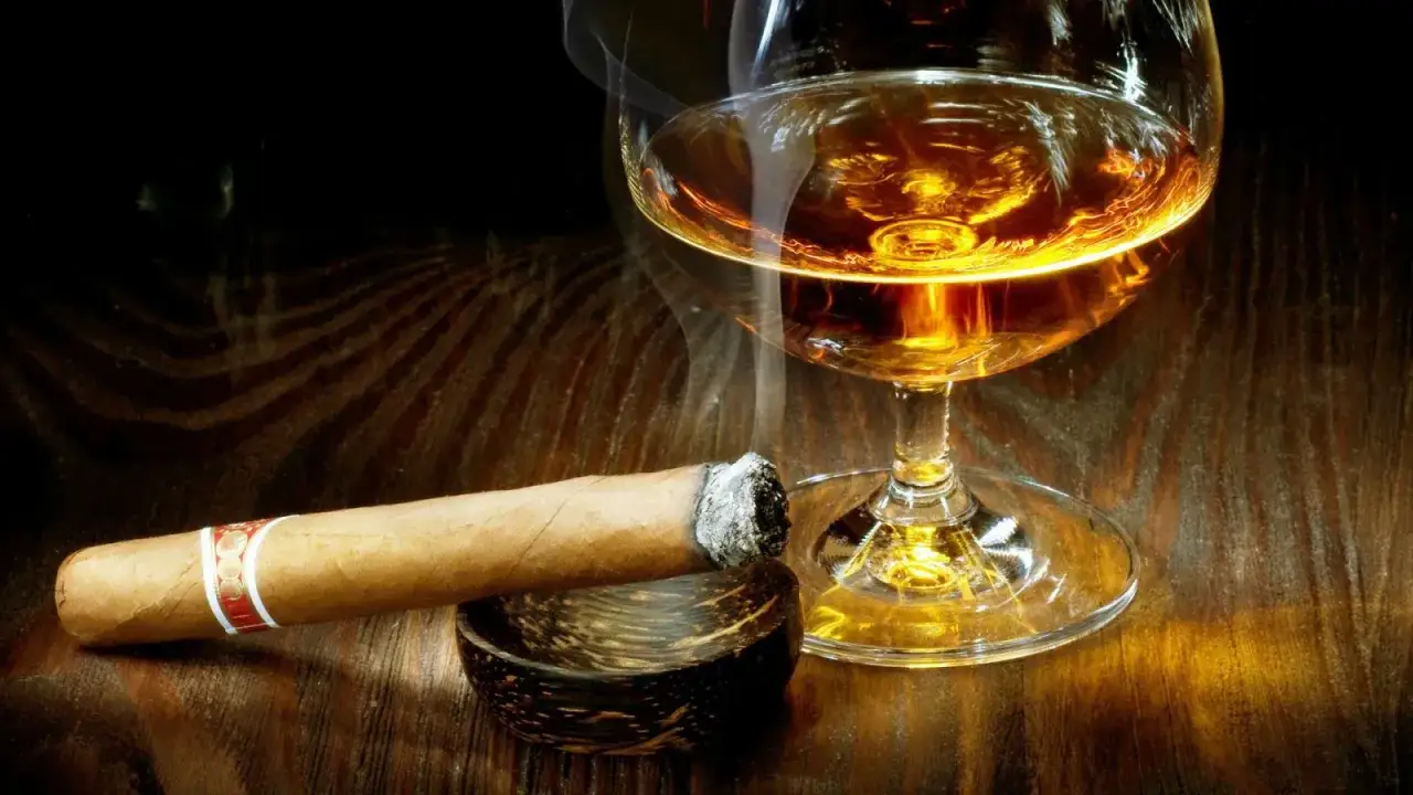 Dymiące cygaro i kieliszek bursztynowej whisky. Czy whisky jest zdrowe? To pytanie pozostaje otwarte, gdy patrzymy na ten obraz.
