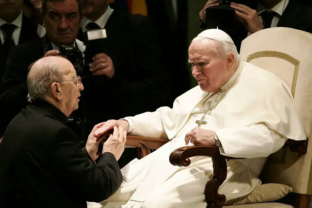 Papież Jan Paweł II w białej szacie, klęczący mężczyzna w czerni. W tle widać fotografów. To obraz duchowej siły, jakiej doświadczają legiony Chrystusa.