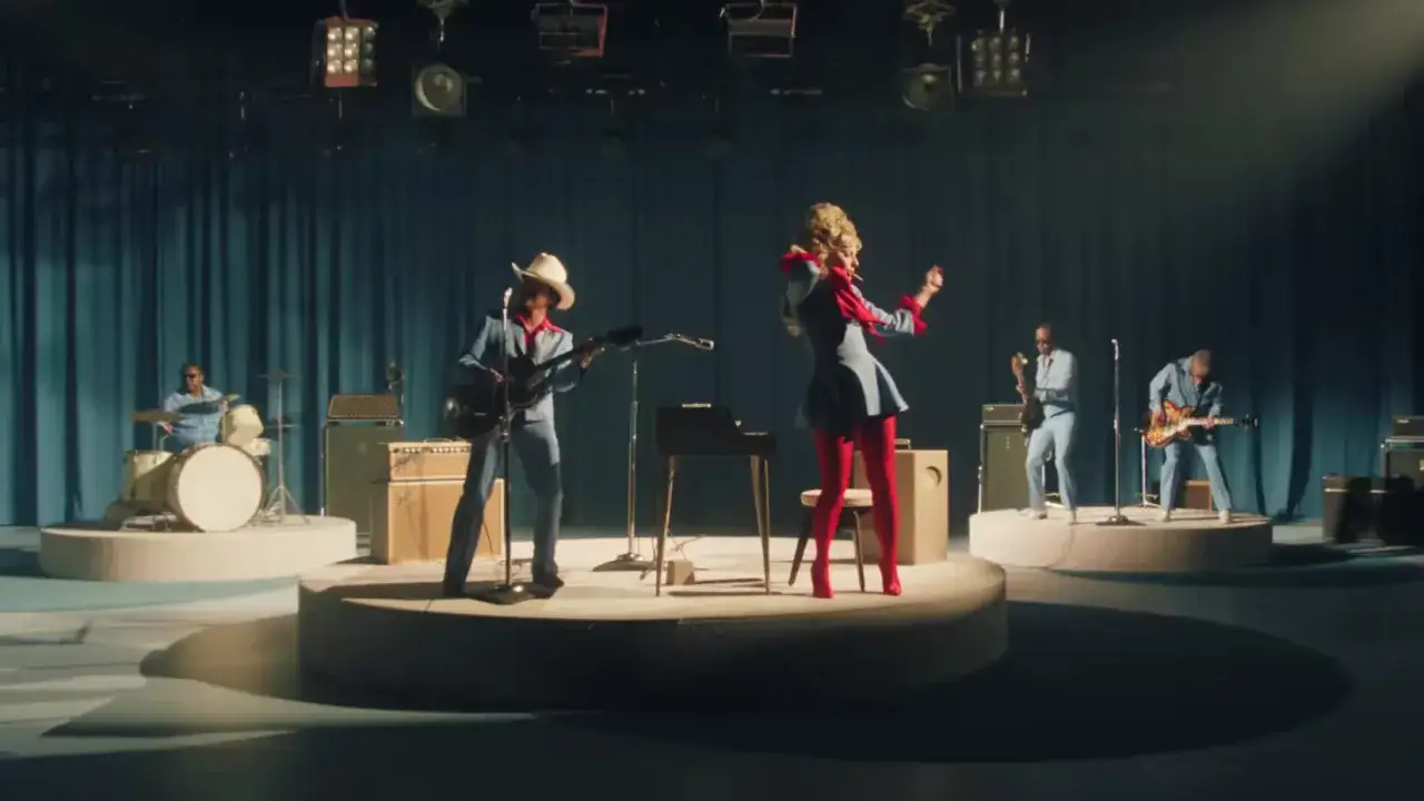 Lady Gaga w czerwonych rajstopach i niebieskiej sukience tańczy na scenie, podczas gdy zespół gra. Tekst "die with a smile" może być inspiracją do tej sceny.