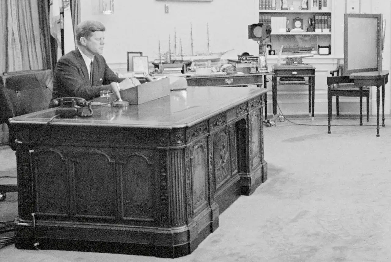 John F. Kennedy am Schreibtisch im Oval Office. Hinter ihm ein Modellschiff, das an die HMS Resolute erinnert, die dem Schreibtisch seinen Namen gab.