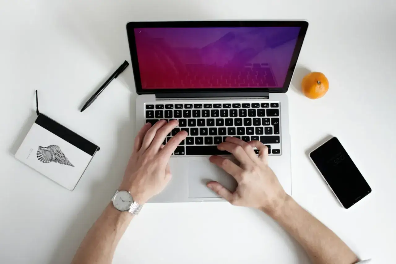 Ręce piszą na laptopie, obok notatnik, długopis, mandarynka i telefon. Idealne do pracy zdalnej, gdy liczy się czas pracy baterii.