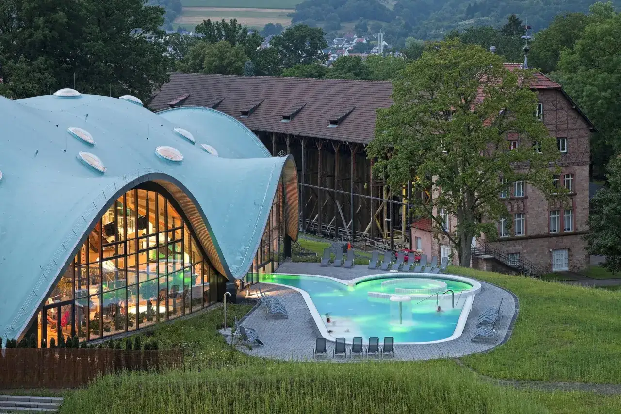 Moderne Therme mit geschwungenem Dach und beleuchtetem Pool, daneben historische Gebäude.