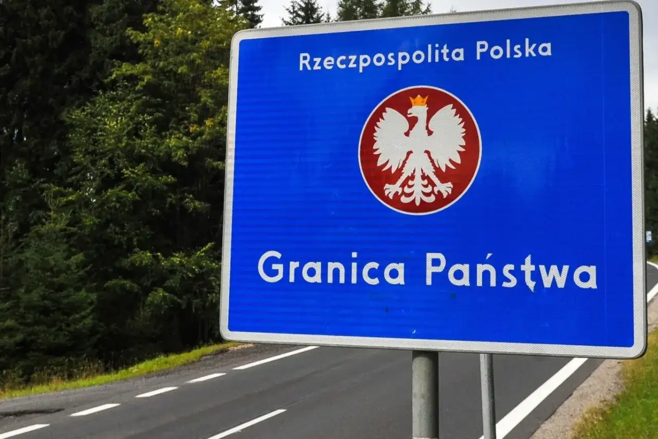 Niebieski znak z orłem i napisem "Rzeczpospolita Polska" oraz "Granica Państwa" stoi przy drodze otoczonej lasem.