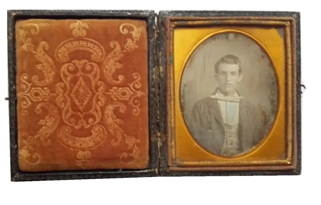Otwarta szkatułka z aksamitną wyściółką i portretem młodego mężczyzny na wyblakłej **daguerreotypii**.