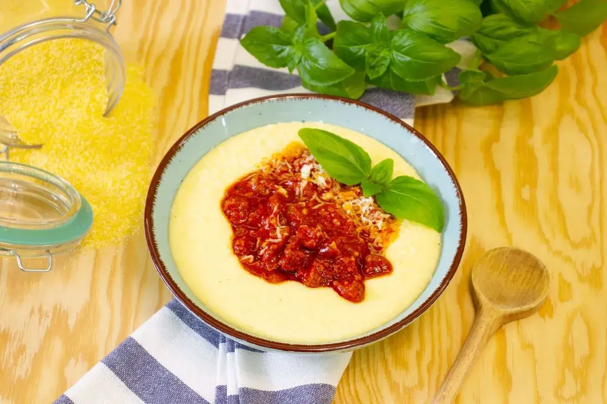 Aksamitna polenta z sosem pomidorowym i parmezanem, ozdobiona listkami bazylii. Obok słoik z mąką kukurydzianą.
