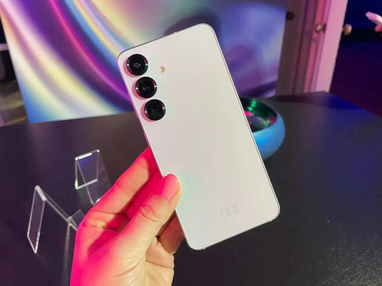 Biały smartfon z trzema aparatami trzymany w dłoni na tle kolorowego, abstrakcyjnego wzoru.