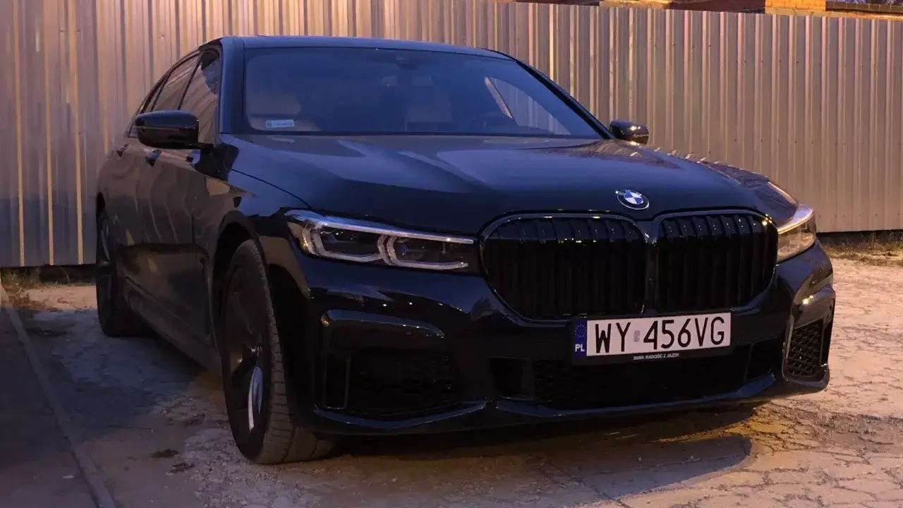 Eleganckie, czarne BMW 750i z charakterystycznym grillem i nowoczesnymi światłami, zaparkowane przy metalowym ogrodzeniu.