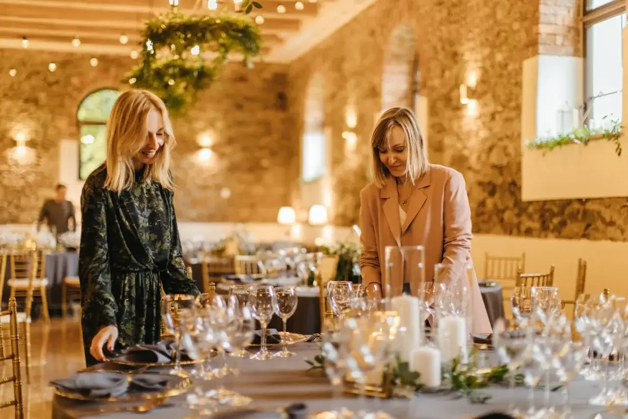 Dwie kobiety przygotowują salę na przyjęcie. Stół nakryty jest eleganckimi naczyniami, tworząc idealny scenariusz wesela.