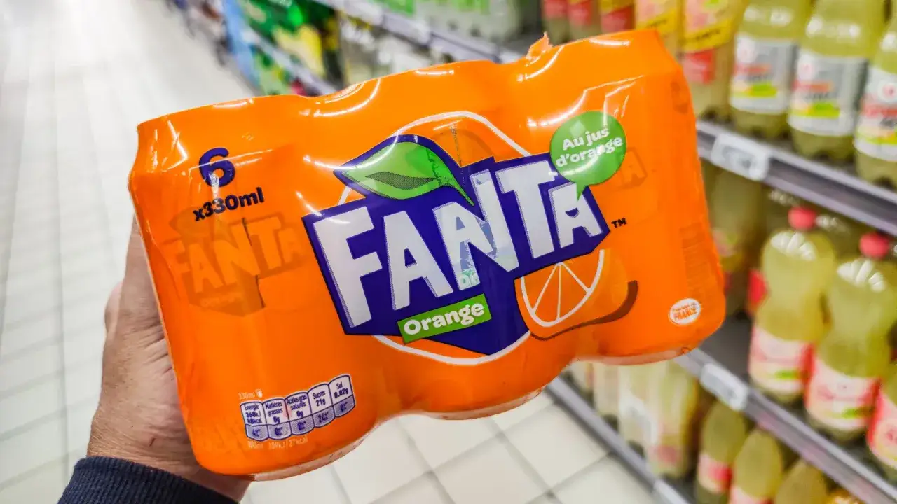 Opakowanie 6 puszek Fanty Orange w ręce, na tle sklepowych półek z napojami.