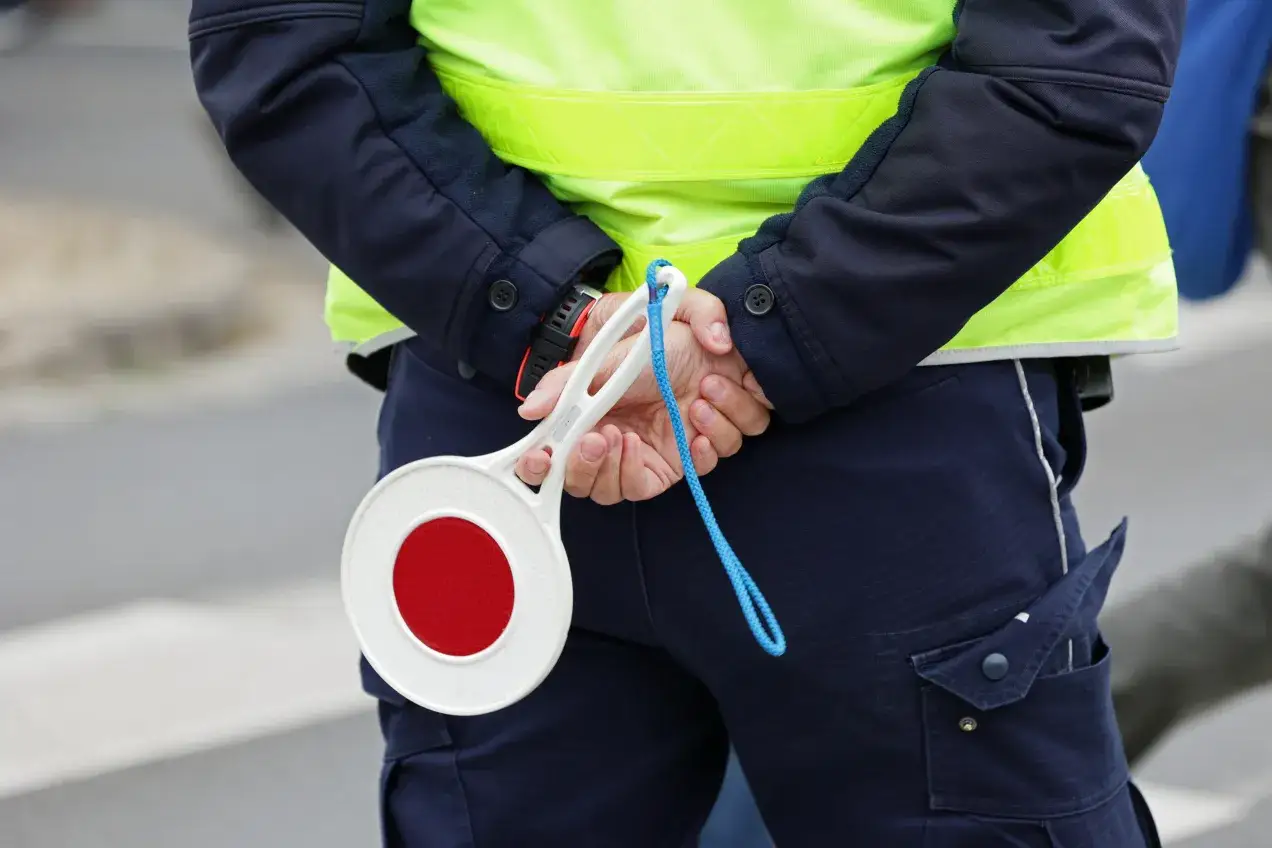 Policjant w kamizelce odblaskowej trzyma za plecami lizak do zatrzymywania pojazdów.