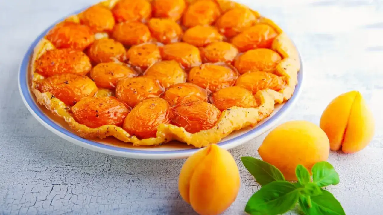 Tarte aux abricots caramélisés sur un fond de tarte croustillant, avec des abricots frais et des feuilles de basilic sur le côté.