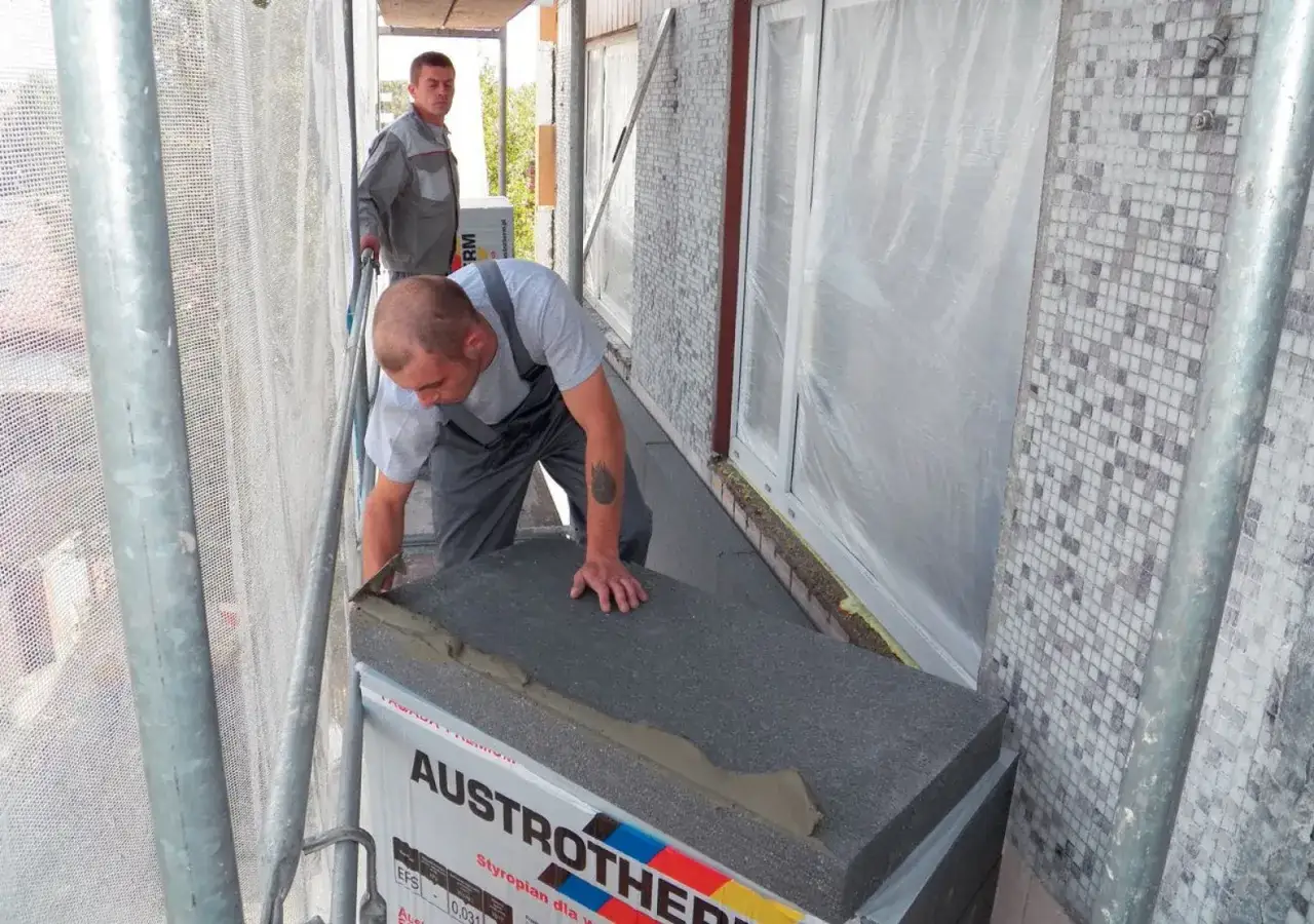 Budowlaniec układa styropian pod wylewkę na balkonie. Obok stoi drugi pracownik.