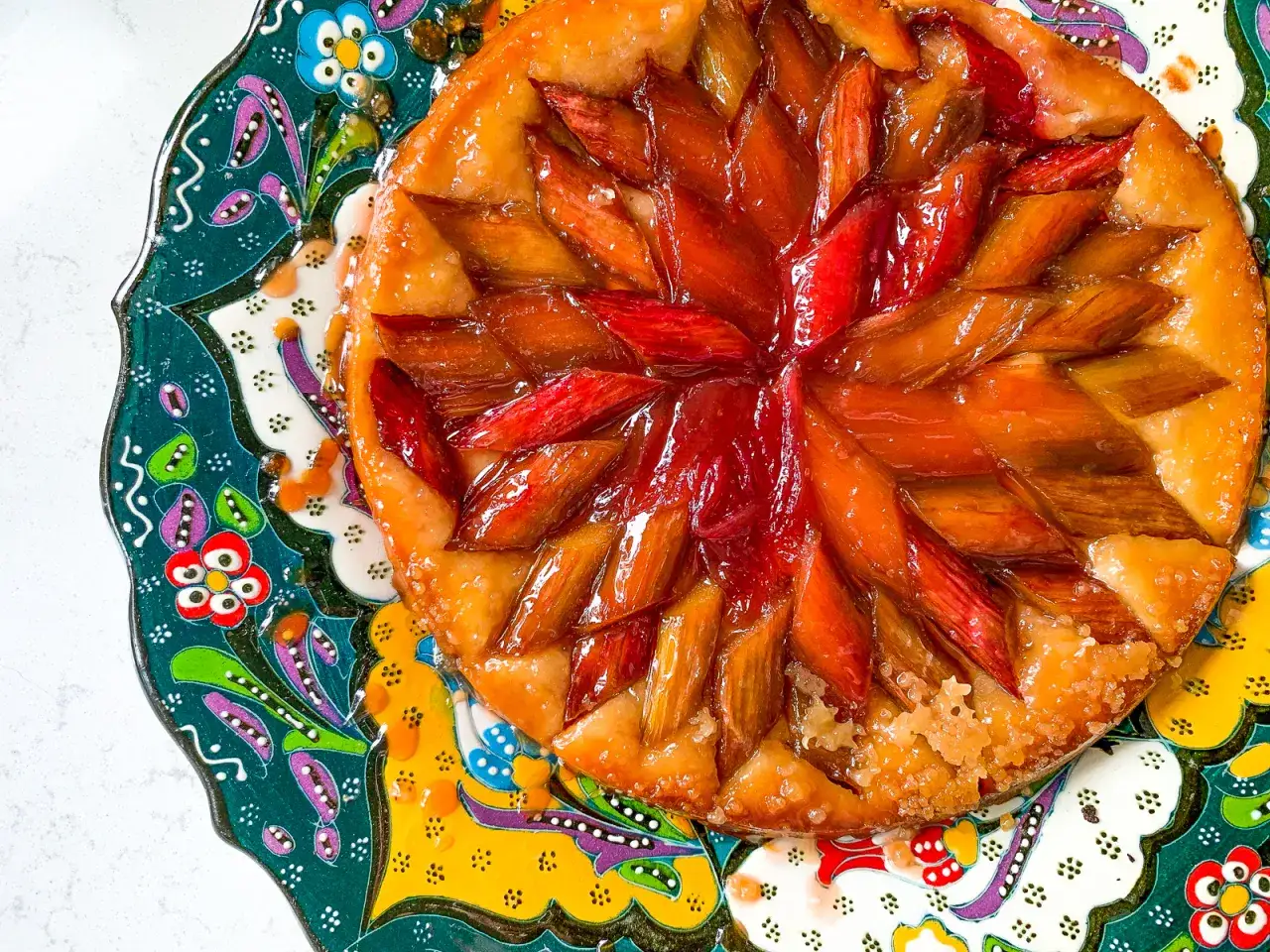 Owoce pod kruszonką, czyli tarta tatin z rabarbarem, błyszcząca od karmelu, na kolorowym, zdobionym talerzu.
