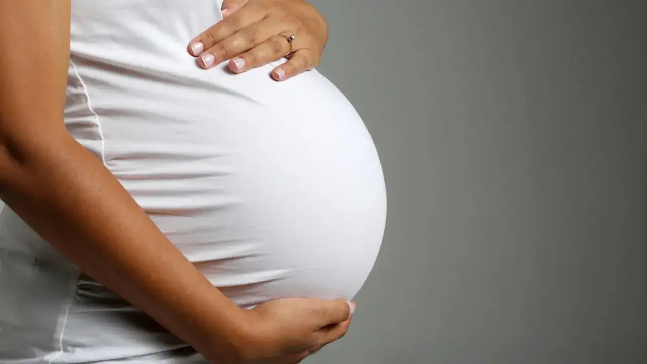 Brzuch ciężarnej kobiety w białej bluzce, dłonie delikatnie go obejmują. Zbliża się poród, może już wkrótce pojawi się czop śluzowy.