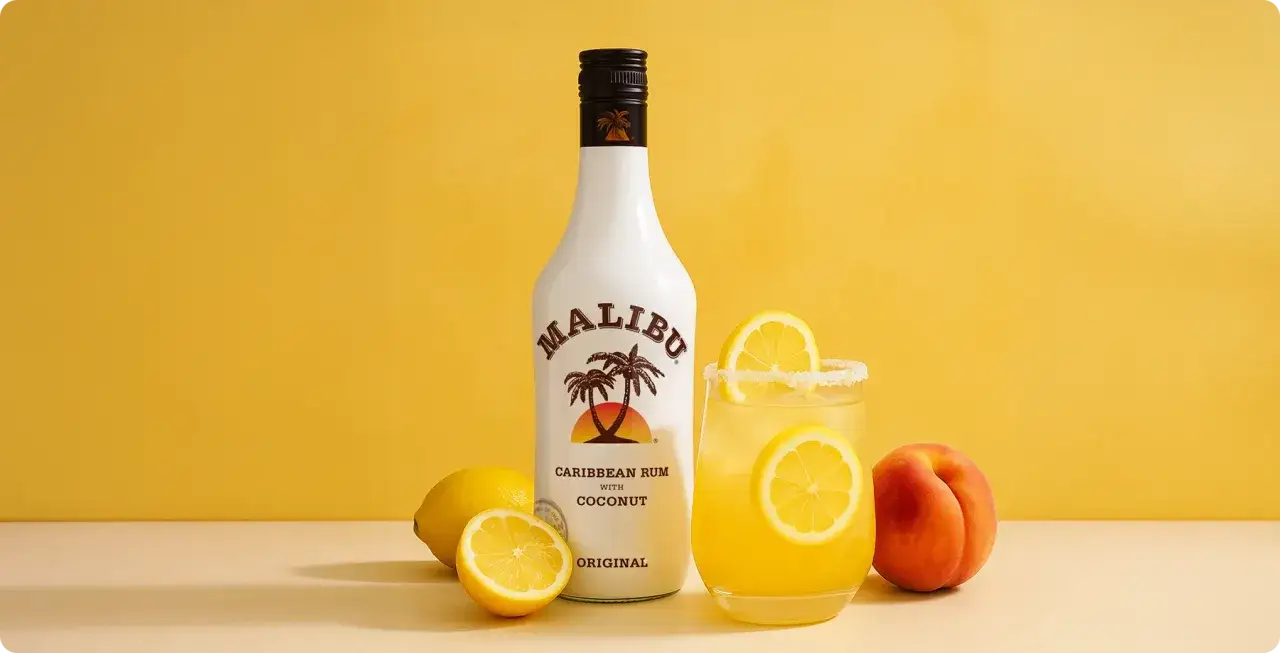 Malibu z mlekiem czy Malibu z sokiem ananasowym? Obok butelki białego rumu Malibu i drinka z cytryną stoi brzoskwinia.