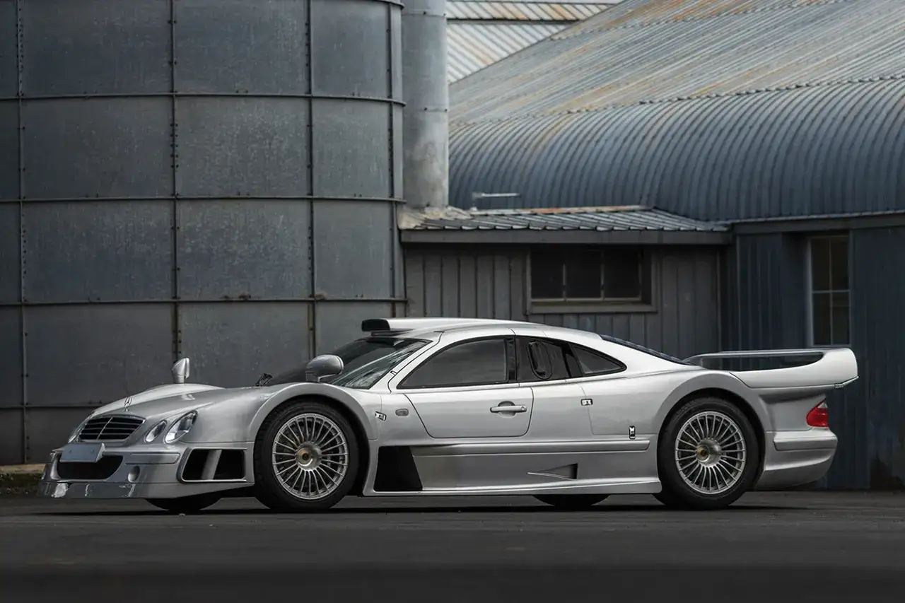 Srebrny Mercedes CLK GTR, ikona wyścigów, zaparkowany przed industrialnymi budynkami.