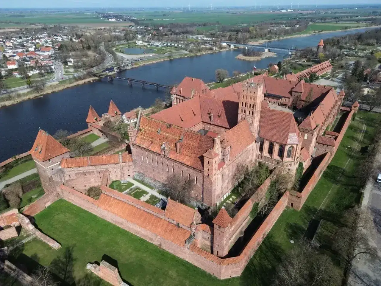 Potężny zamek krzyżacki, największy zamek w Europie, wznosi się nad rzeką Nogat. Czerwona cegła i liczne wieże tworzą imponujący widok.