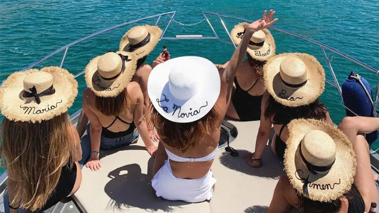 Grupo de amigas en un barco celebrando unas divertidas ideas despedida de soltera. Llevan sombreros de paja con nombres bordados, uno dice "La Novia".