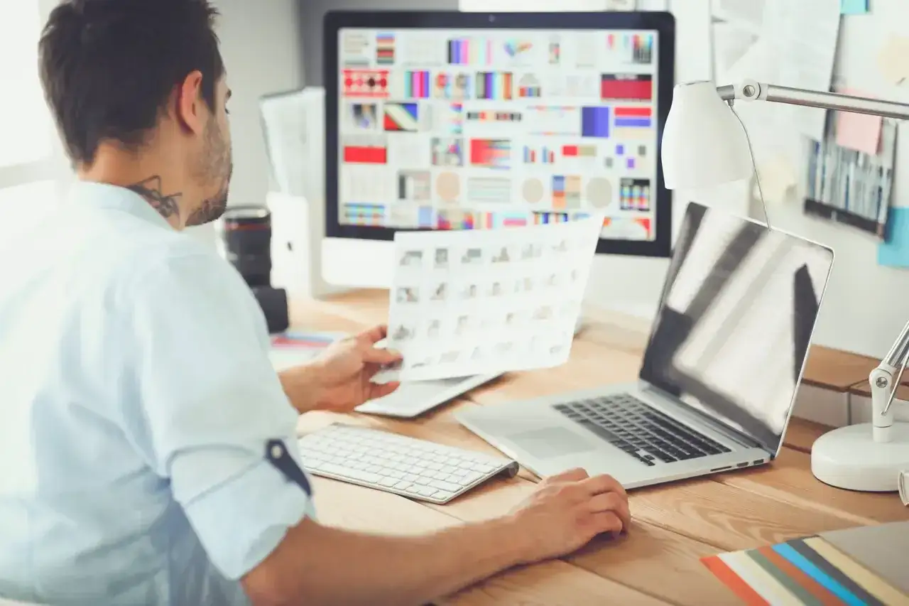 Mężczyzna, jako ux designer, analizuje wydruki przy biurku z komputerem i laptopem.