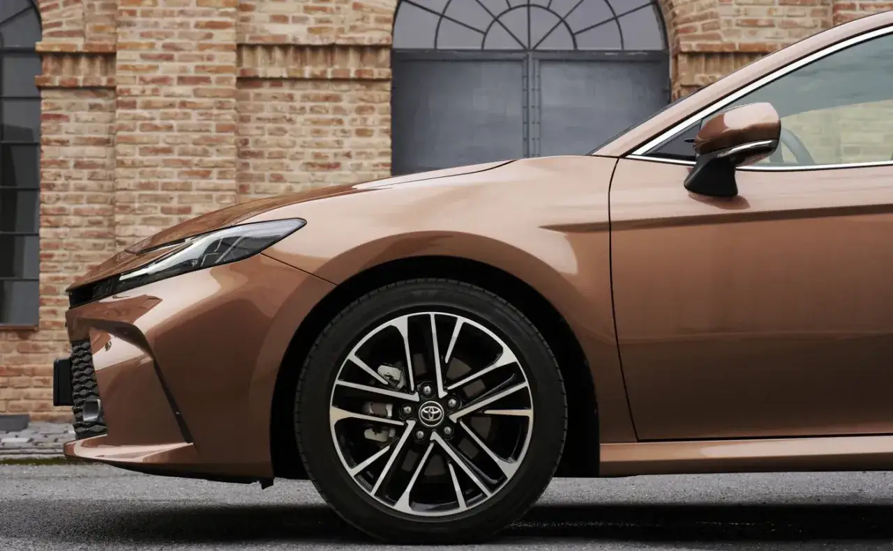 Elegancka, brązowa Toyota Camry 2024 z nowymi światłami i felgami, zaparkowana przed ceglanym budynkiem.