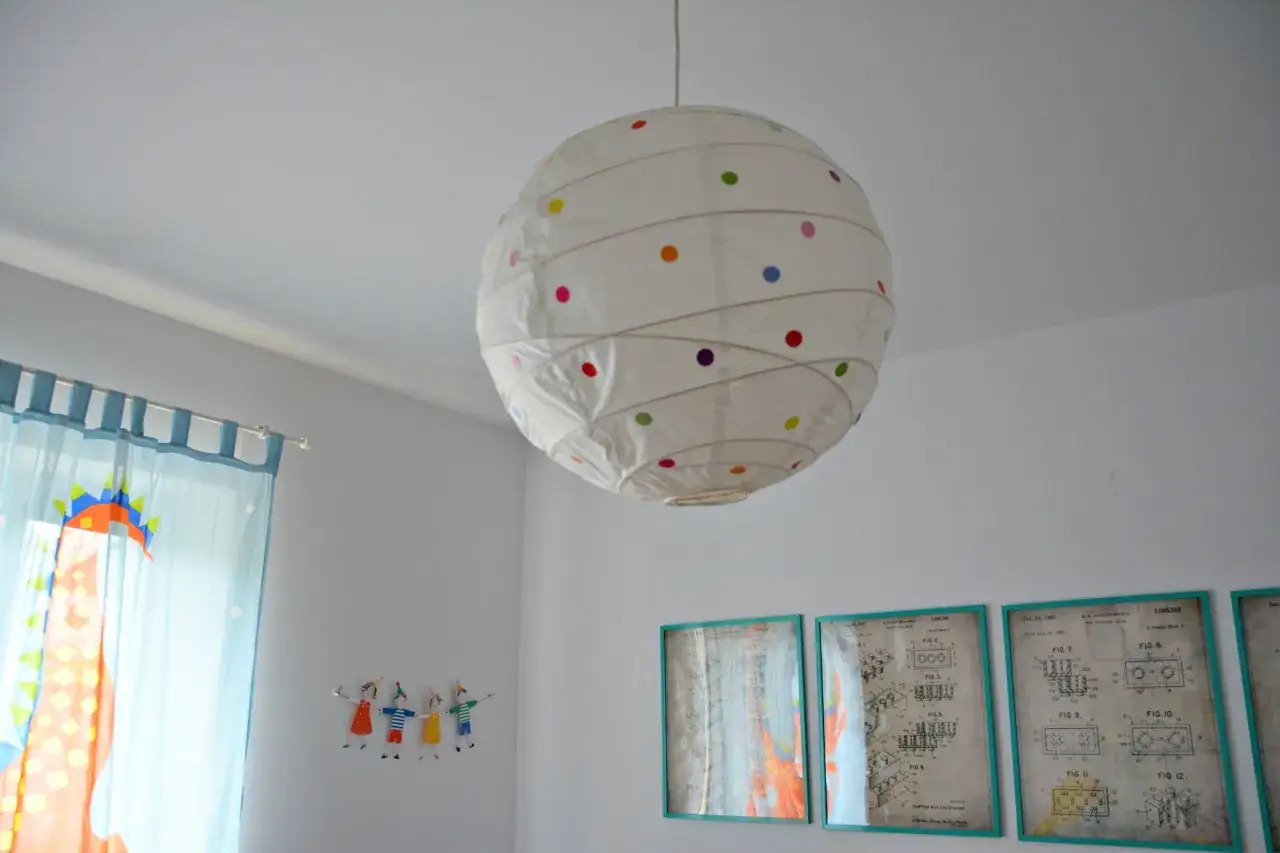 Jak odnowić papierową lampę? Biała lampa wisząca w kropki, obok niebieskich zasłon z dinozaurem i rysunków dzieci.