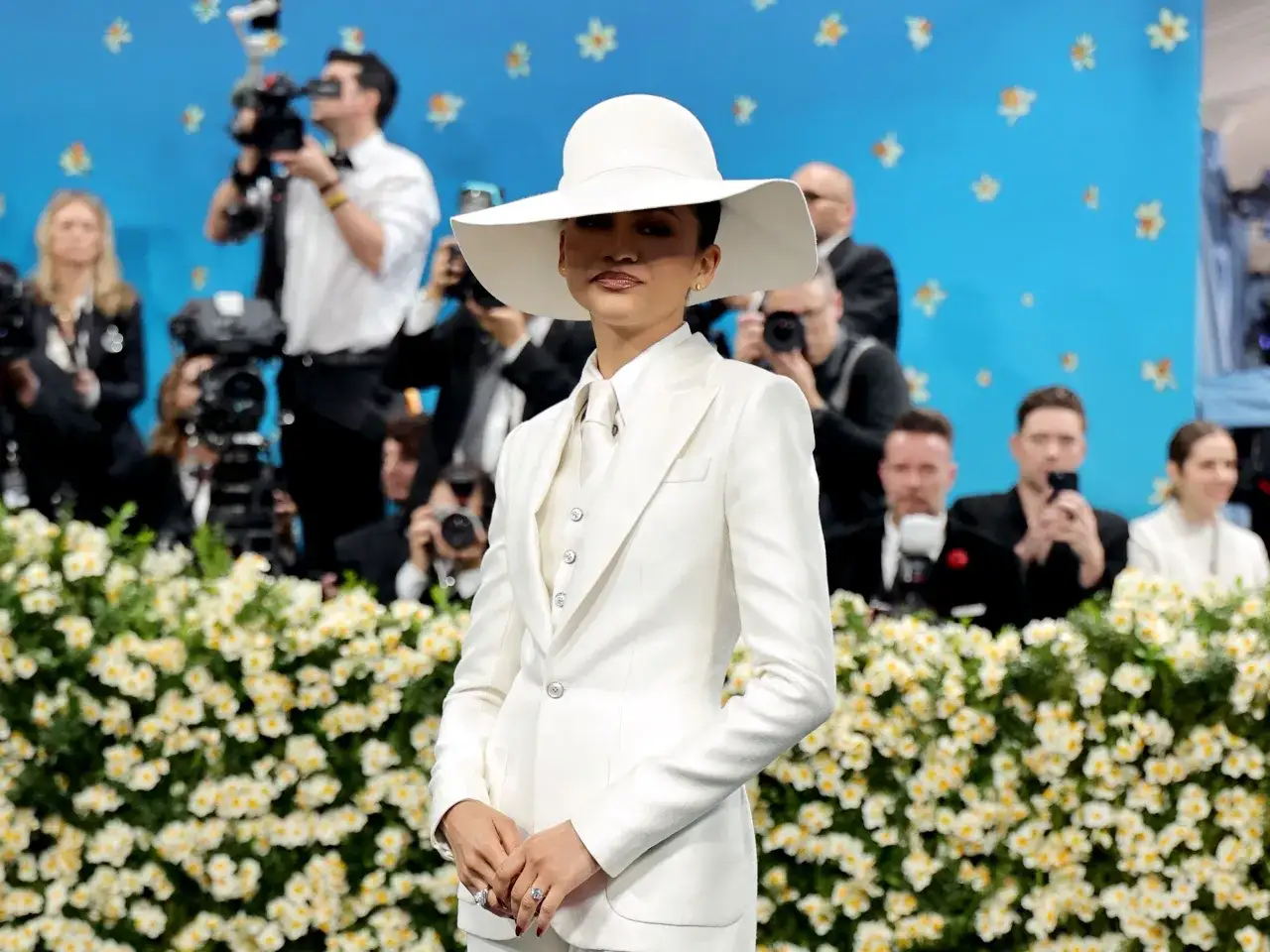 Zendaya, deslumbrante con traje blanco y sombrero, posa ante cámaras y flores.