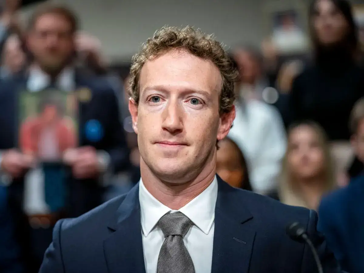 Mark Zuckerberg im Anzug, im Hintergrund unscharfe Menschen und ein Foto.