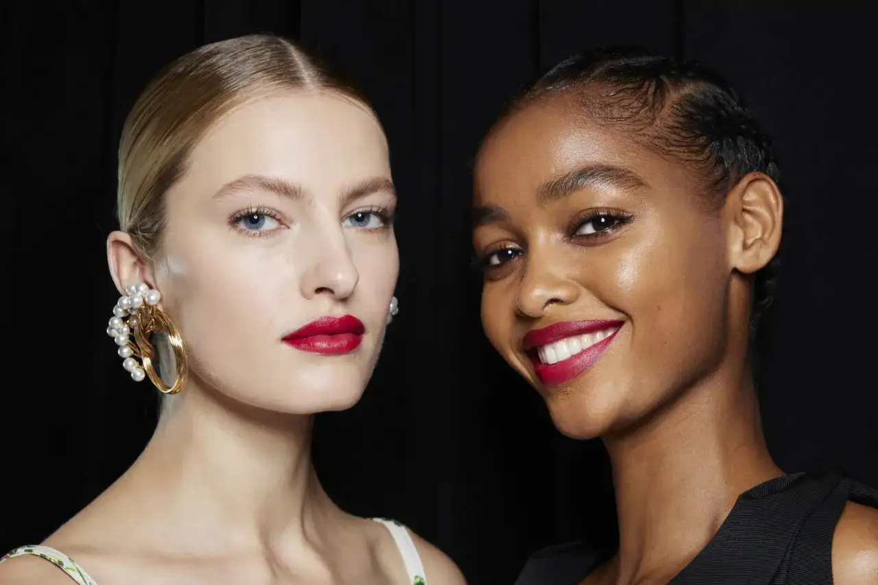 Dos modelos lucen un maquillaje para boda con labios rojos vibrantes y piel radiante. Una lleva un pendiente de perlas, la otra sonríe.