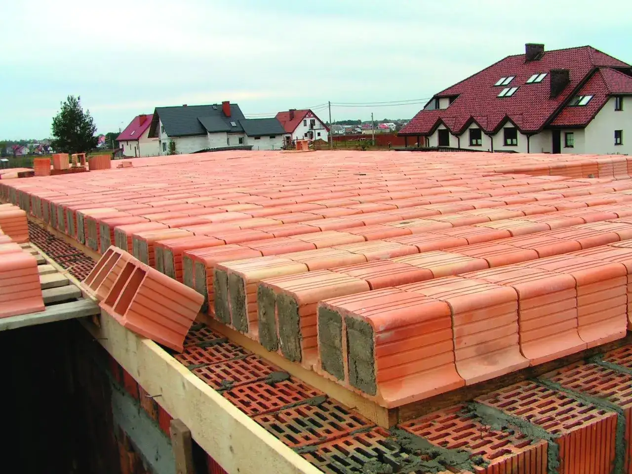 Budowa stropu ackermana. Czerwone pustaki ceramiczne ułożone w rzędach, gotowe do zalania betonem. W tle domy z czerwonymi dachami.