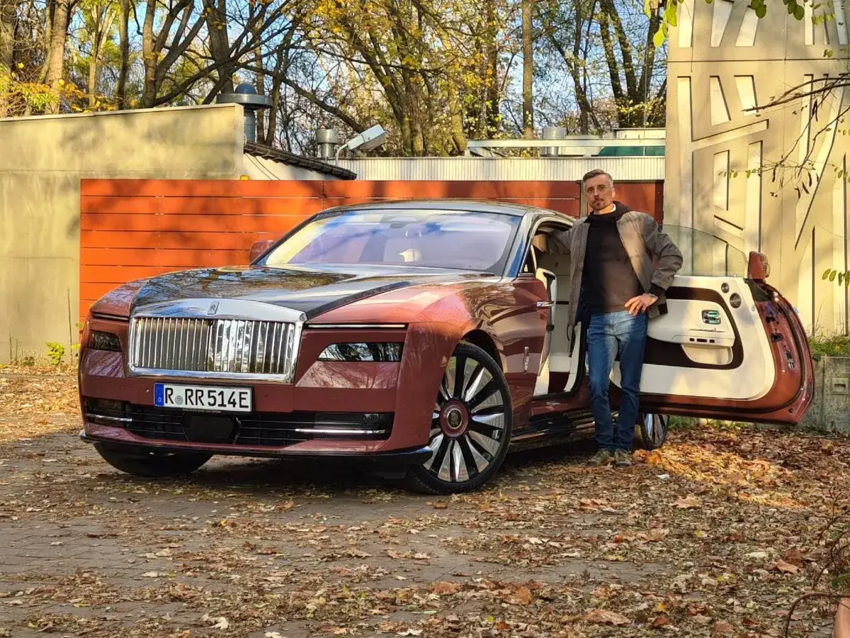 Elegancki mężczyzna obok luksusowego Rolls-Royce'a elektrycznego w kolorze bordowym. Jesienne liście wokół.