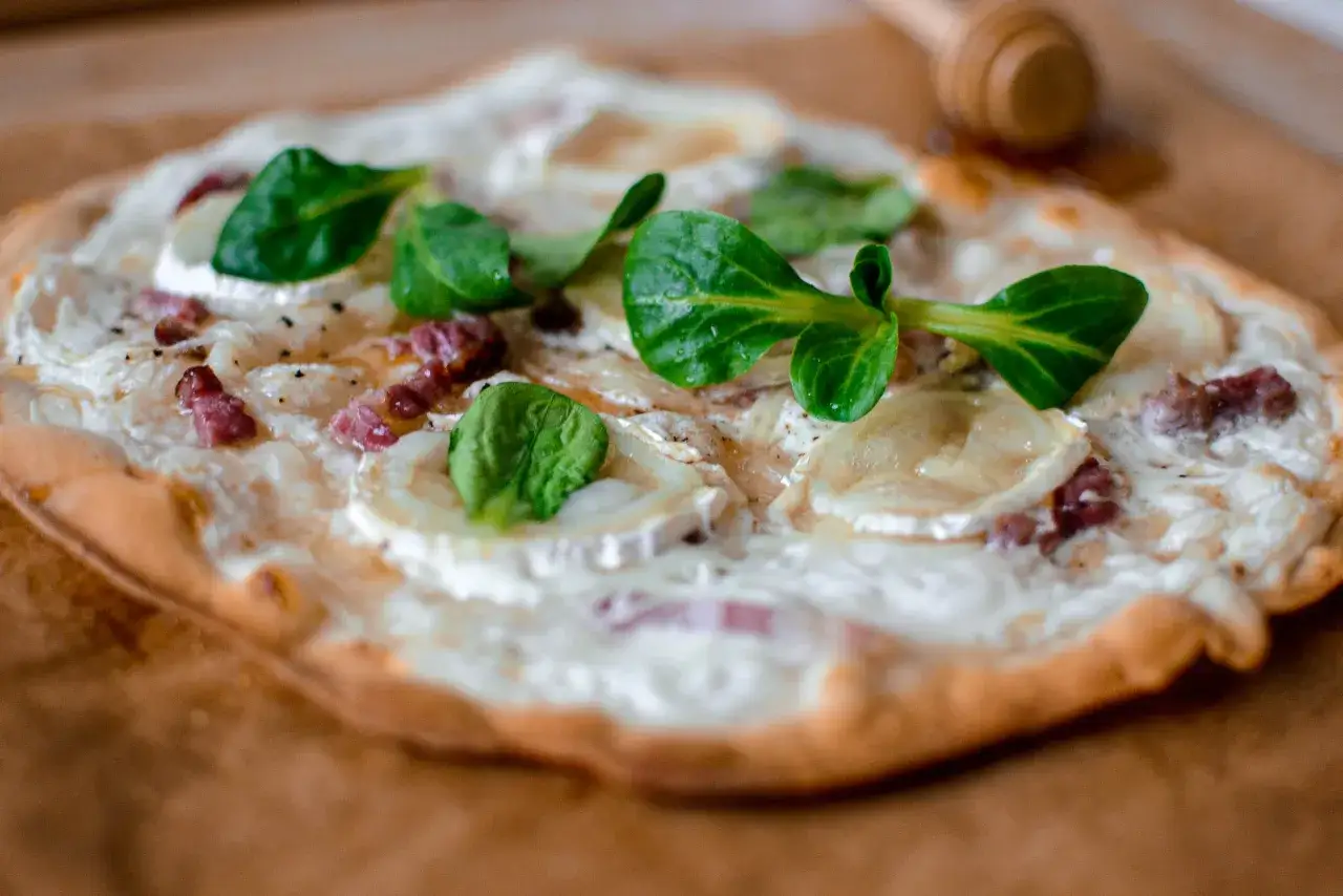 Flammekueche alsace garnie de lardons, fromage de chèvre et jeunes pousses d'épinards sur une pâte fine et croustillante.