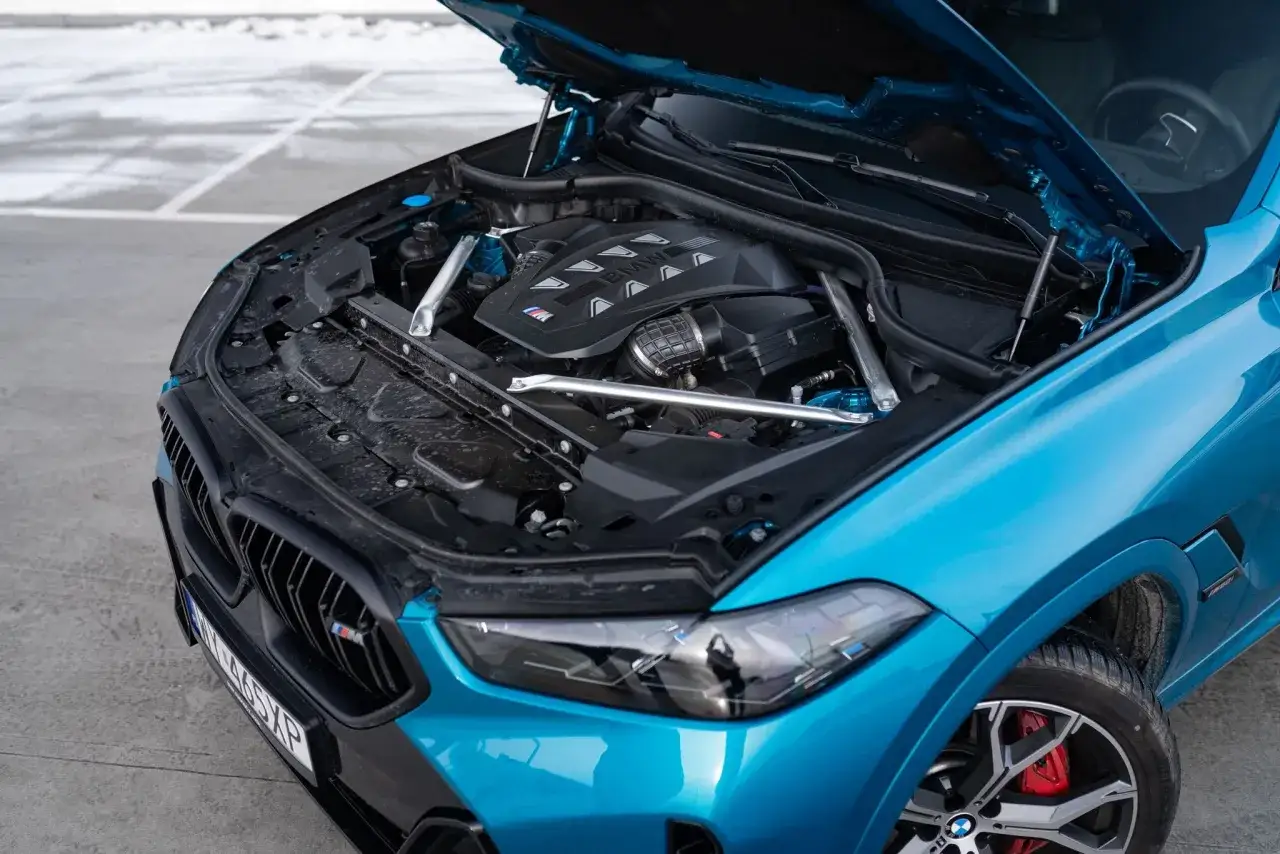 Błękitne x6 BMW z otwartą maską, odsłaniającą potężny silnik M. Widoczne detale podkreślają sportowy charakter auta.