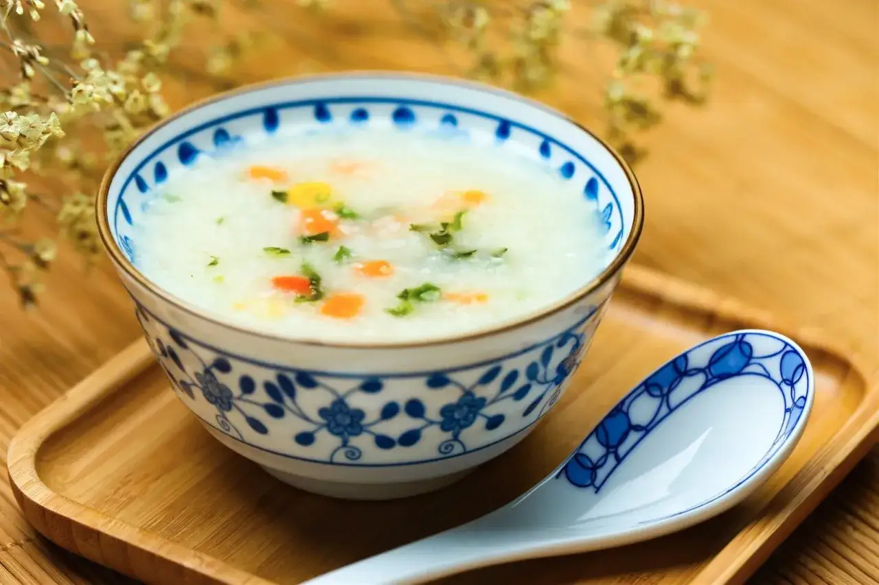 Miseczka z congee, ryżową potrawą z warzywami, obok łyżki na drewnianej tacce.