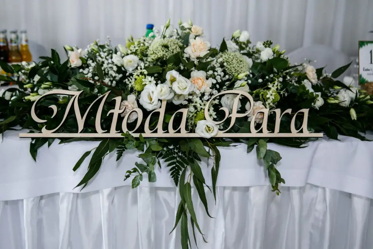 Drewniany napis "Młoda Para" na stole weselnym, ozdobionym bujną zielenią i białymi kwiatami.