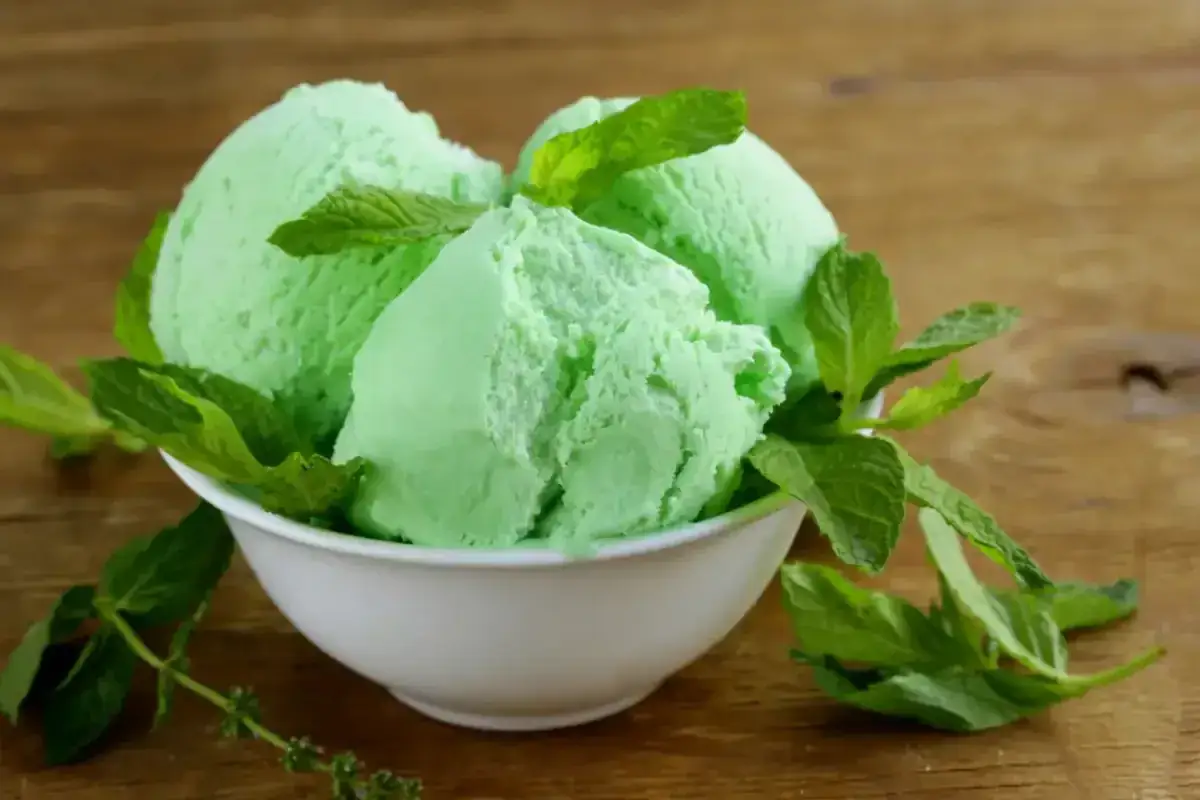 Trois boules de glace verte menthe, rappelant la saveur de la glace Get 27, garnies de feuilles de menthe fraîche dans un bol blanc sur fond de bois.