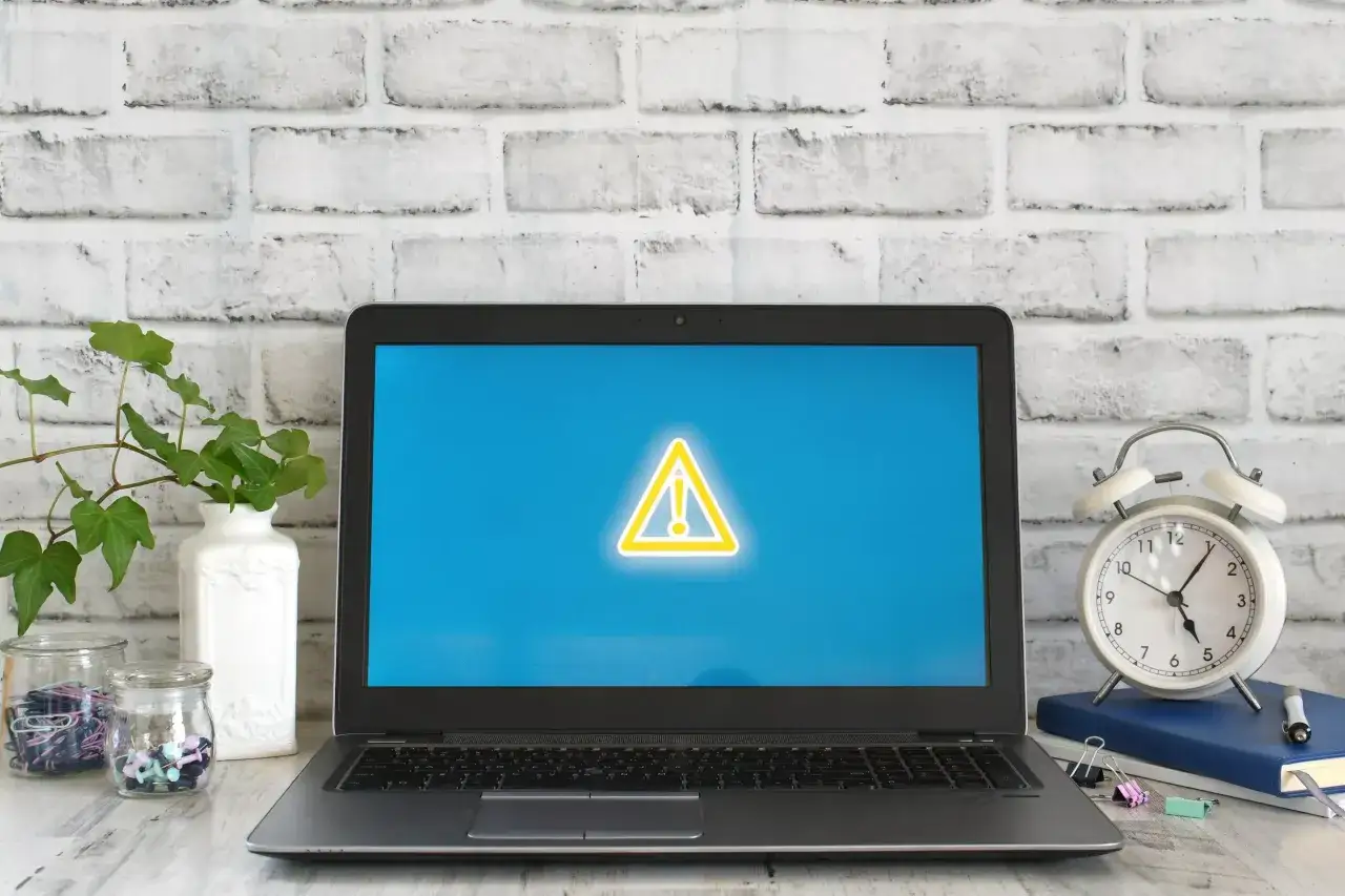 Laptop z symbolem ostrzeżenia na ekranie, obok budzik i rośliny.