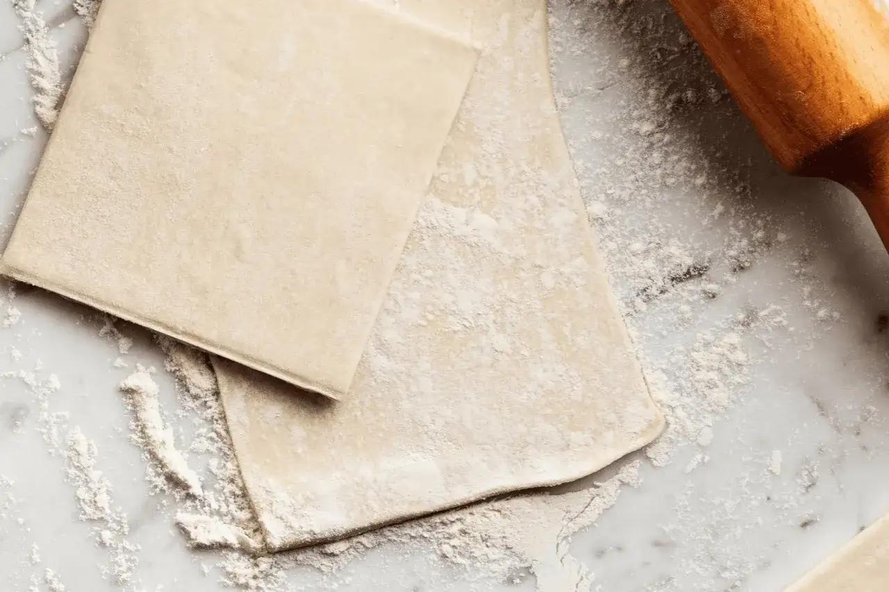 Pâte feuilletée prête à être travaillée, saupoudrée de farine sur un plan de travail en marbre. Un rouleau à pâtisserie en bois est visible sur la droite.