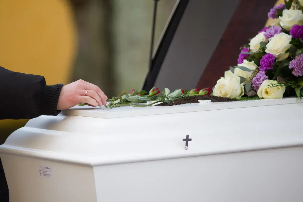 Dłoń z obrączką kładzie róże na białej trumnie, ozdobionej bukietem kwiatów i krzyżem.