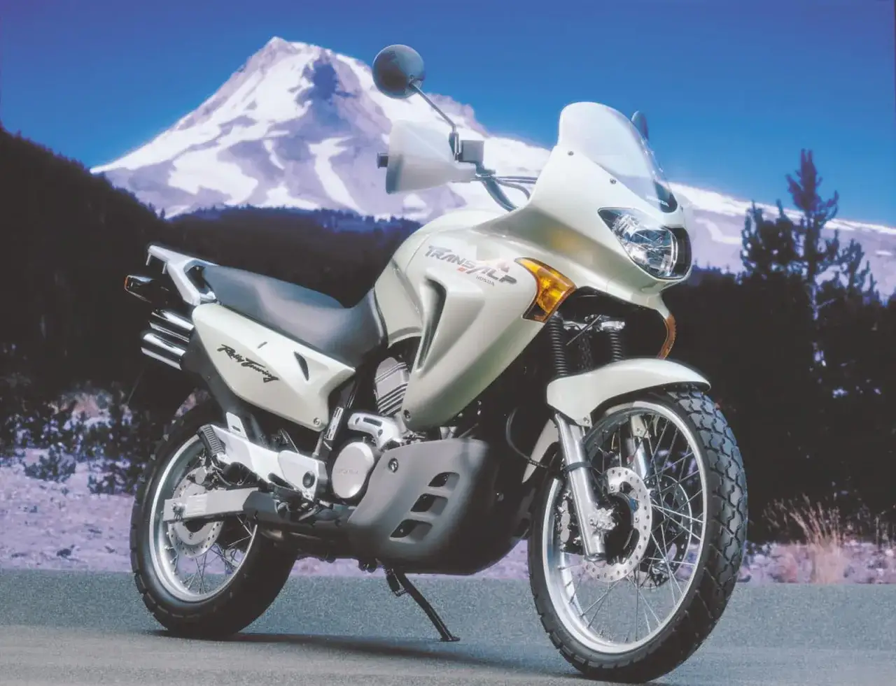 Srebrny motocykl Honda Transalp 650 gotowy na przygodę na tle ośnieżonej góry i lasu.