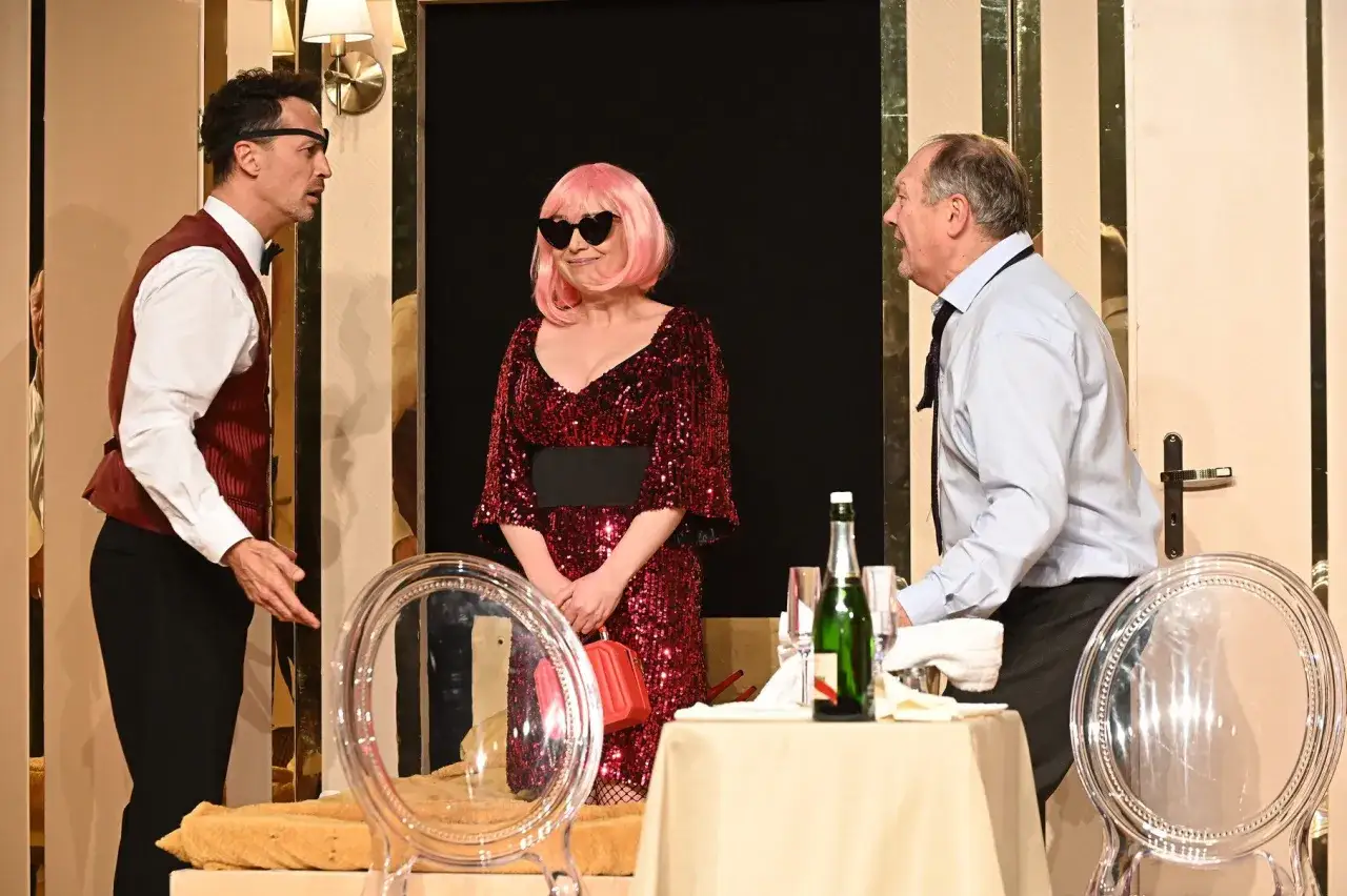 Trzech aktorów na scenie Teatru Capitol Kraków. Kobieta w błyszczącej sukni i peruce, mężczyźni w eleganckich strojach.